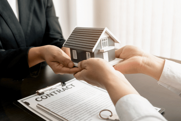 Two people exchange a small model house over a contract on a table, symbolizing a rental or real estate agreement.