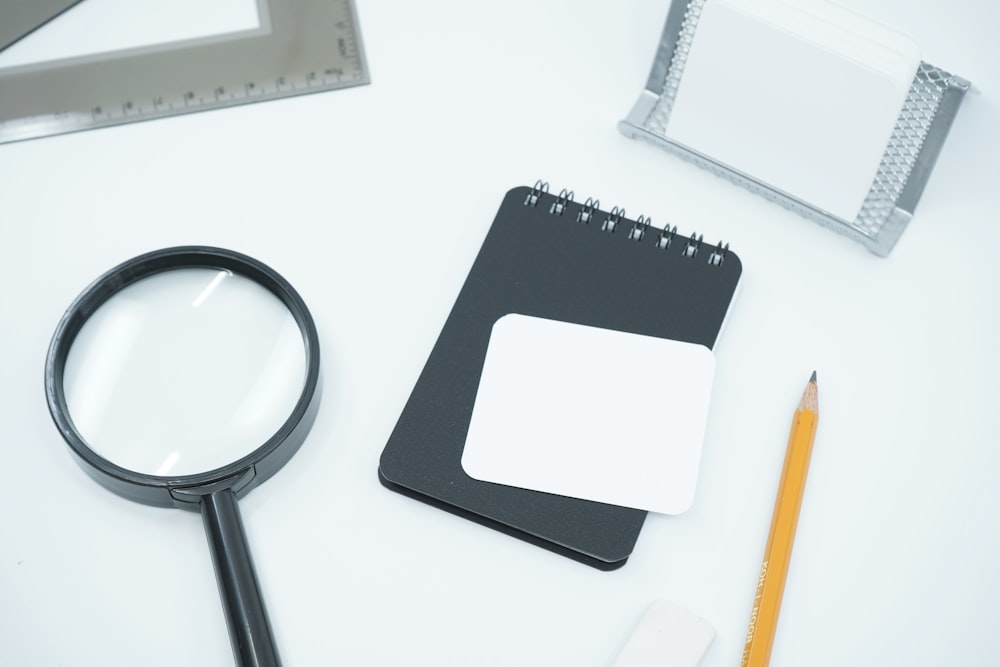Magnifying glass, black notebook, pencil, and ruler on white desk representing investigation and research tools