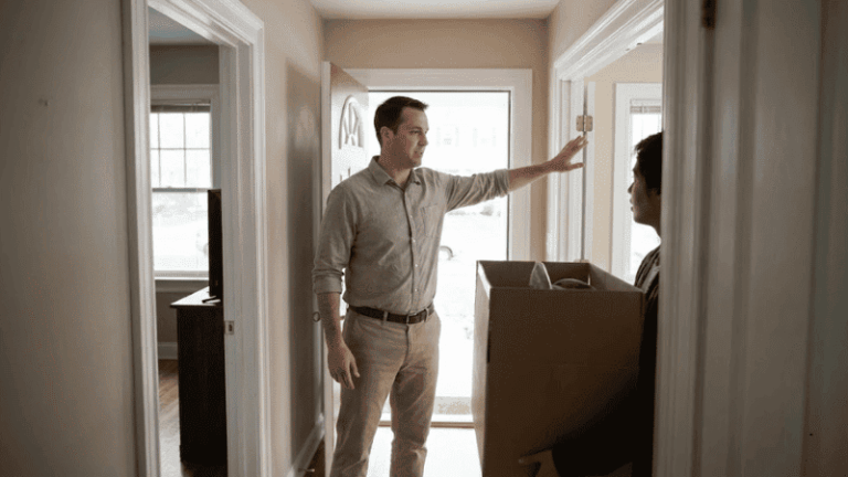 A man stands in the doorway of his home speaking to another person holding a large cardboard box, suggesting a move-in or move-out situation.